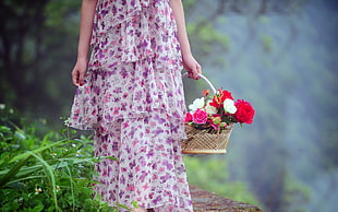 person wearing pink and purple floral dress carrying basket of flower standing on green grass field